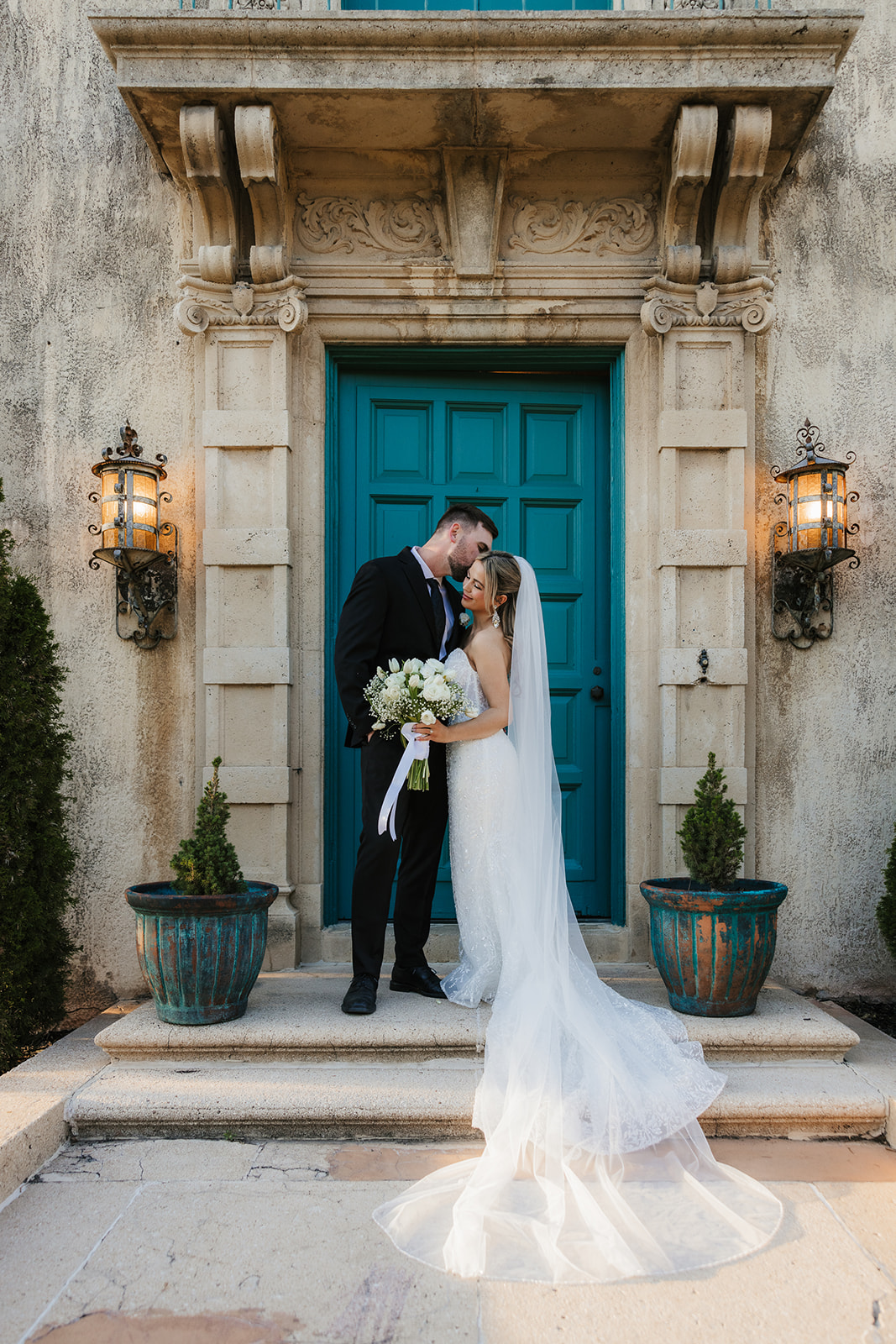 Bride and groom standing on steps of a historic building with blue doors and windows, flanked by tall potted plants. A modern cylindrical structure is visible in the background | Top 6 Historic Mansion Wedding Venues in Tulsa
