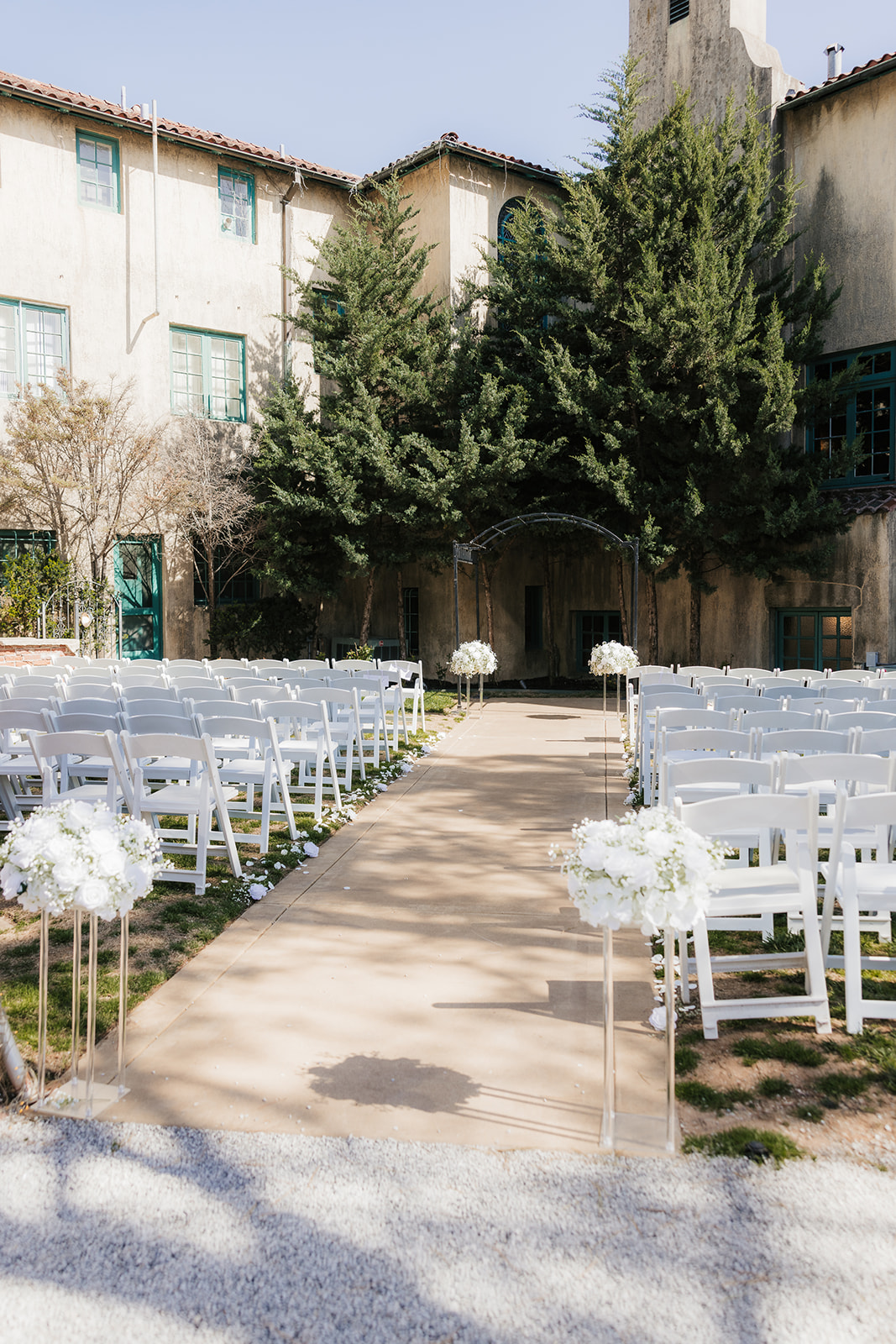 Outdoor wedding ceremony setup with white chairs and floral arrangements lining a central aisle, set against a backdrop of trees and a rustic building. | Top 6 Historic Mansion Wedding Venues in Tulsa