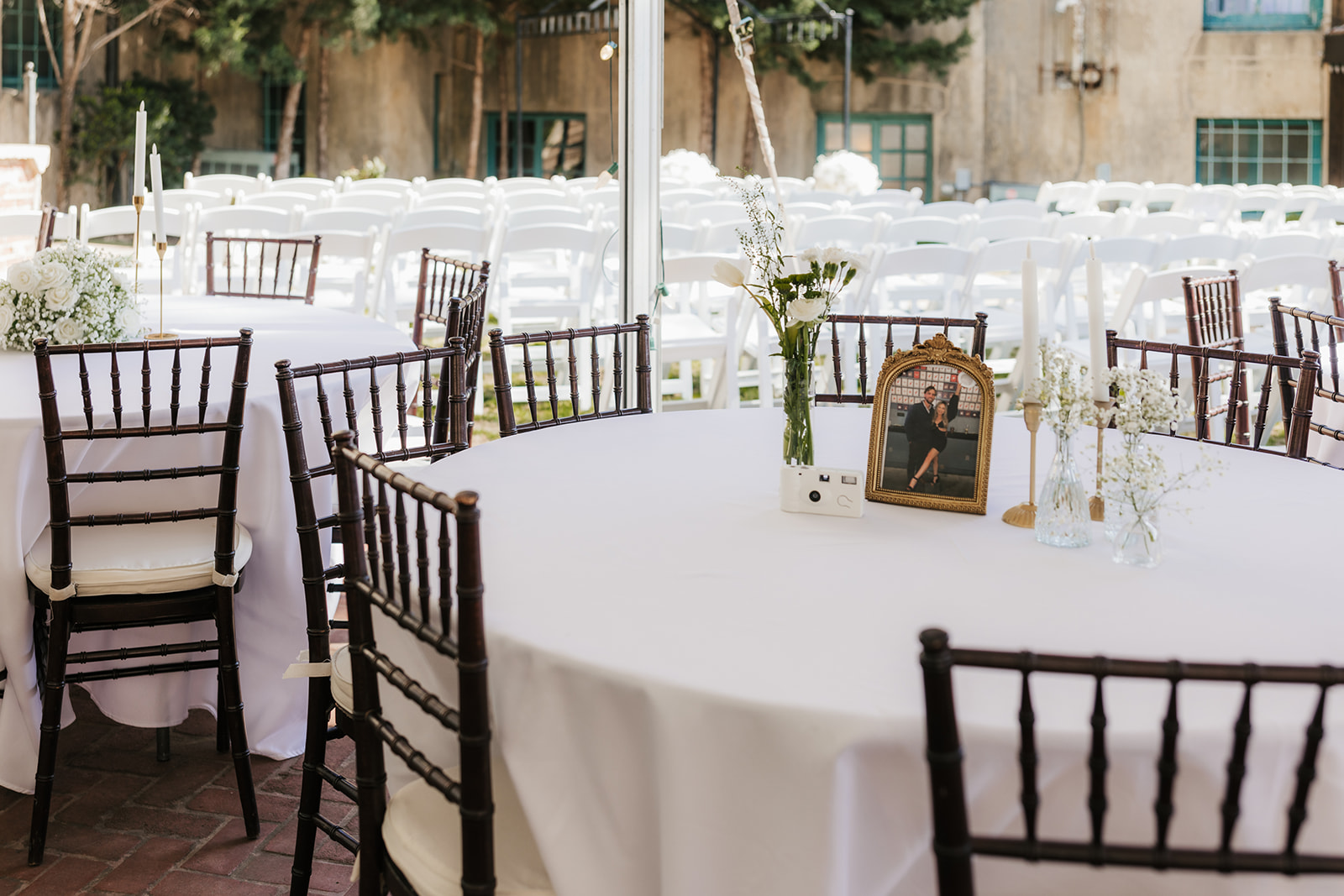 Outdoor wedding reception setup with round tables covered in white cloths. Wooden chairs surround the tables, which have floral centerpieces and a framed photo. Rows of chairs are in the background.