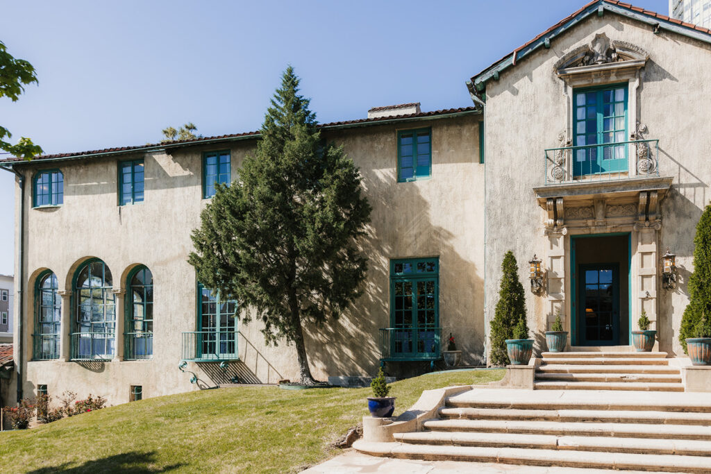 Historic house with stucco exterior, blue-framed windows, and arched glass doors. Steps lead to an entrance with potted plants. A tree is near the facade, surrounded by a grassy lawn at woodway park which is a Historic Mansion Wedding Venues