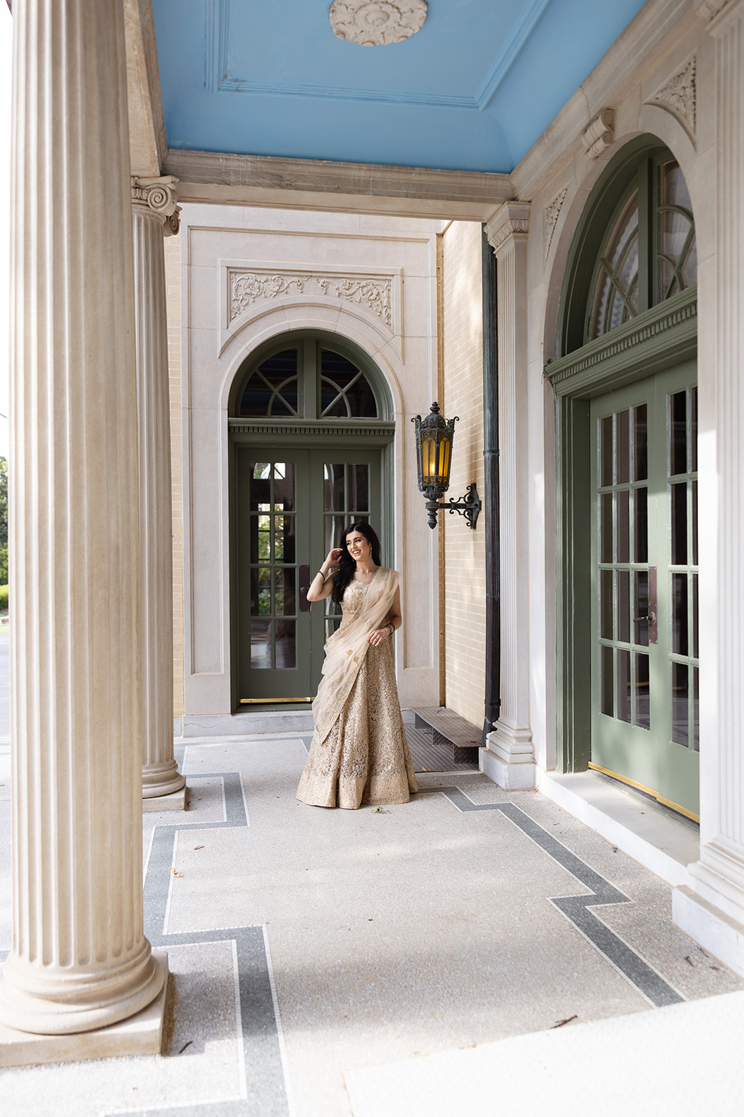 Person wearing a beige outfit stands in a columned porch area with green doors and a blue ceiling.