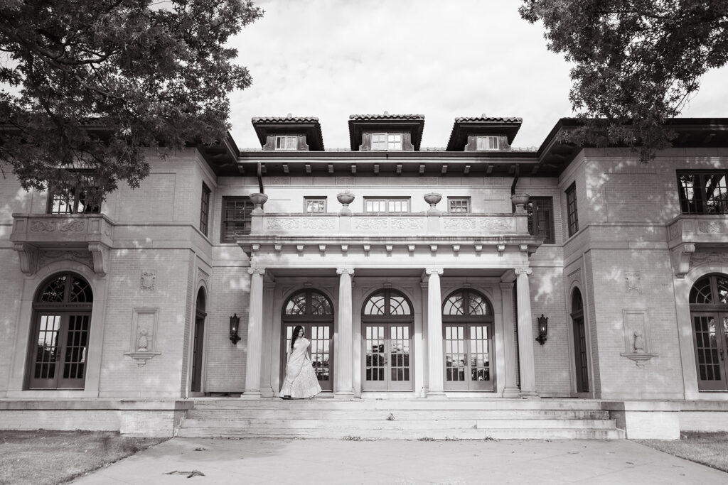 Black and white image of a large, historic building with a bride in a wedding dress standing on the steps. The building features columns, arches, and ornate windows. Trees frame the scene. | Top 6 Historic Mansion Wedding Venues in Tulsa