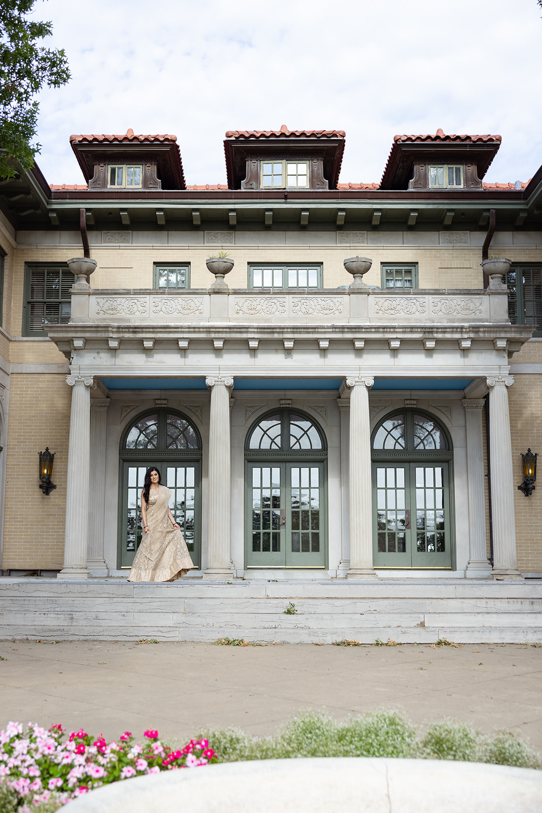 A person in a formal dress stands on the steps of a large building with arched windows and columns, surrounded by greenery.