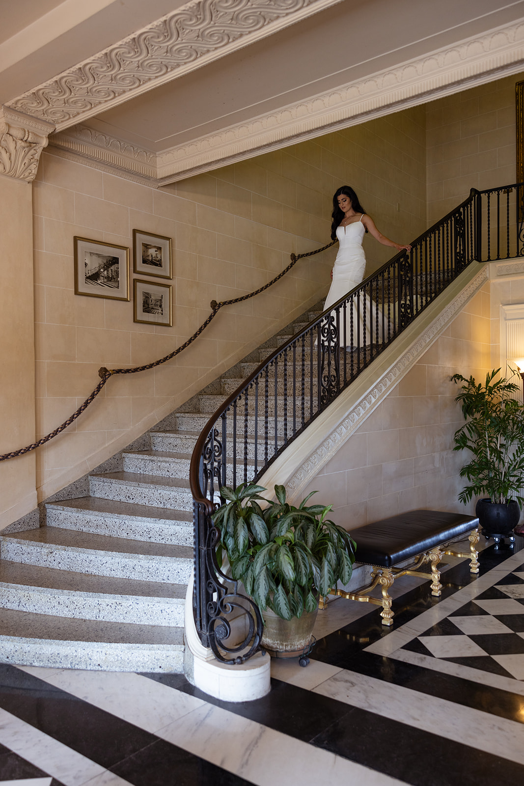 A person in a white dress descends a grand, curved staircase in an elegant hallway with checkered flooring and framed pictures on the wall.