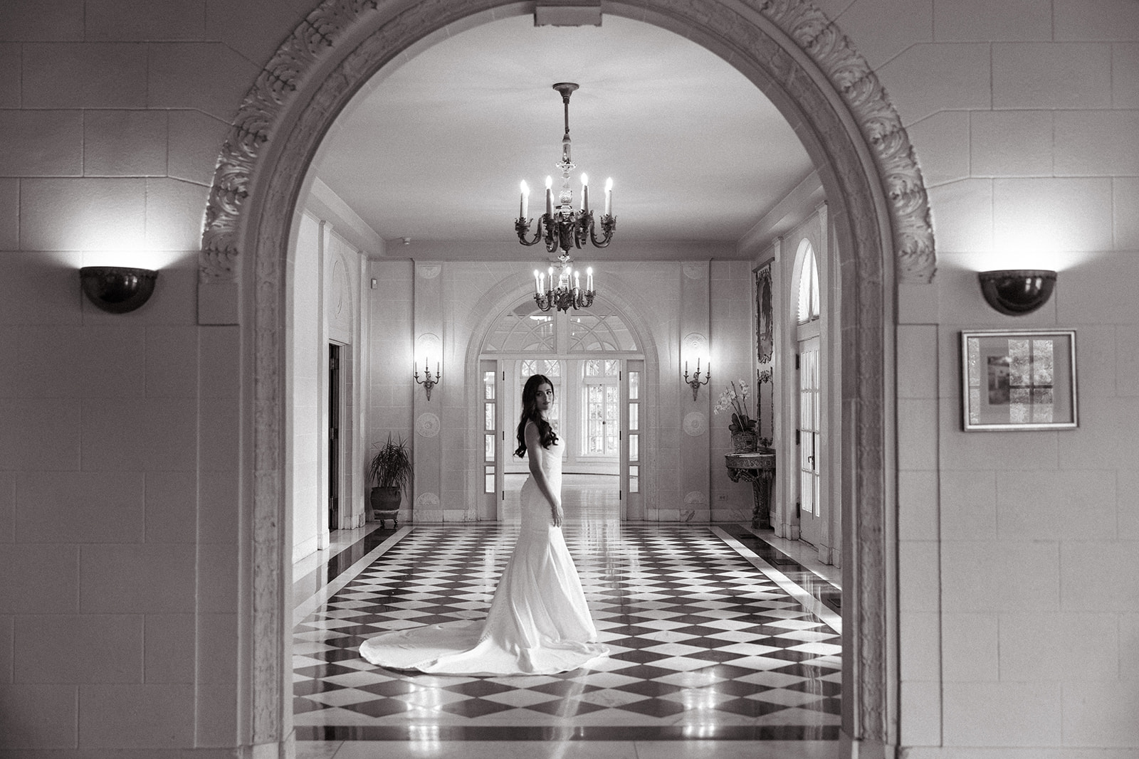   A woman in a long white dress stands in an elegant hallway with checkered flooring and a chandelier above.