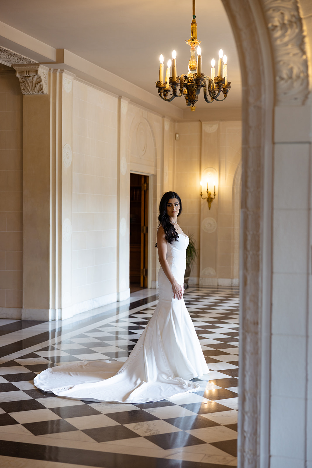 A woman in a long white dress stands in an elegant hallway with checkered flooring and a chandelier above.