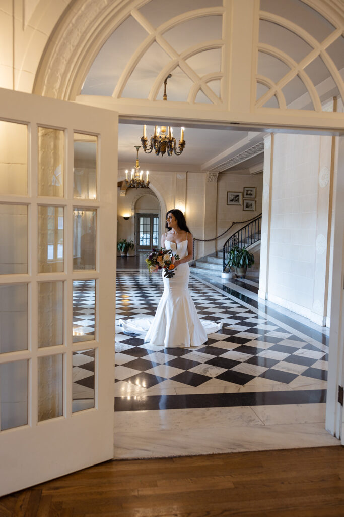 A woman in a long white dress stands in an elegant hallway with checkered flooring and a chandelier above.