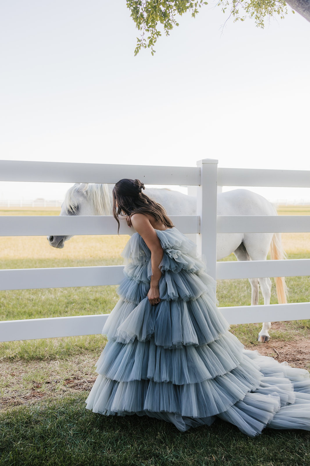 Woman in a flowing layered gown stands by a white fence, reaching towards a white horse at aspen ranch