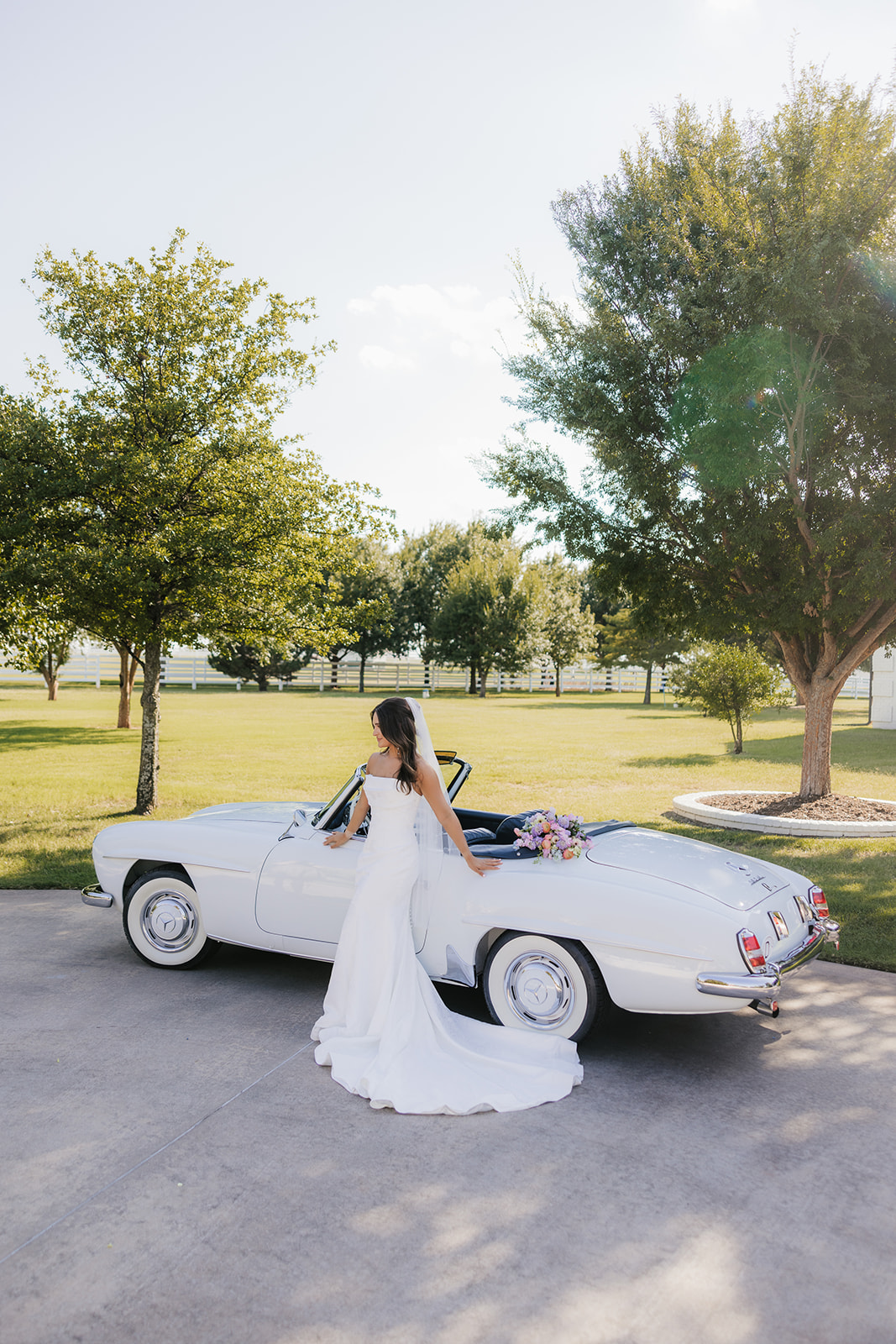 Bride in a white gown stands by a classic white convertible car, holding a bouquet. The setting is an outdoor area with trees and grass under a clear sky at aspen ranch