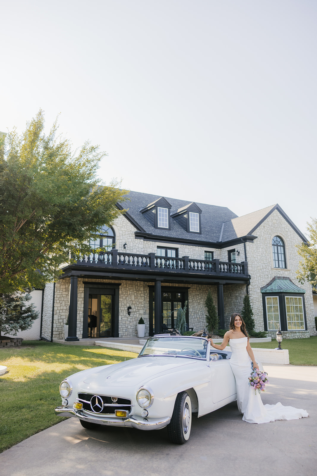 Bride in a white gown stands by a classic white convertible car, holding a bouquet. The setting is an outdoor area with trees and grass under a clear sky at aspen ranch