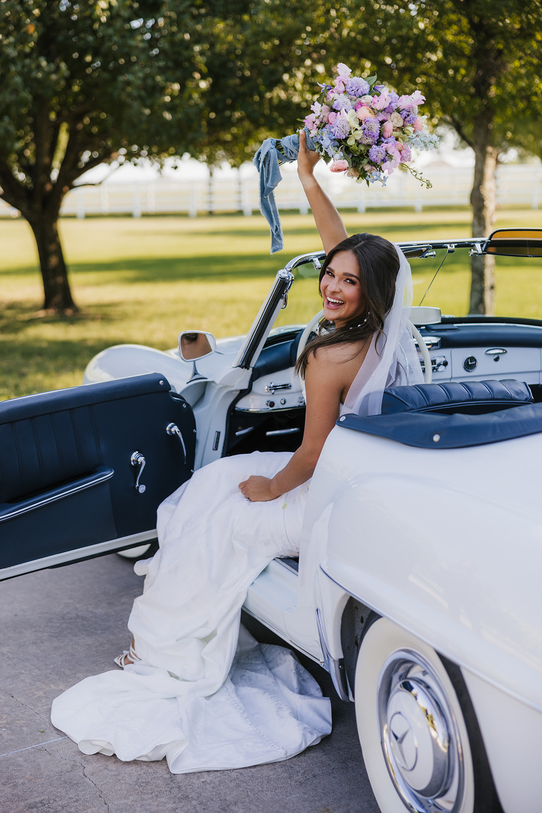 Bride in a white gown stands by a classic white convertible car, holding a bouquet. The setting is an outdoor area with trees and grass under a clear sky at aspen ranch