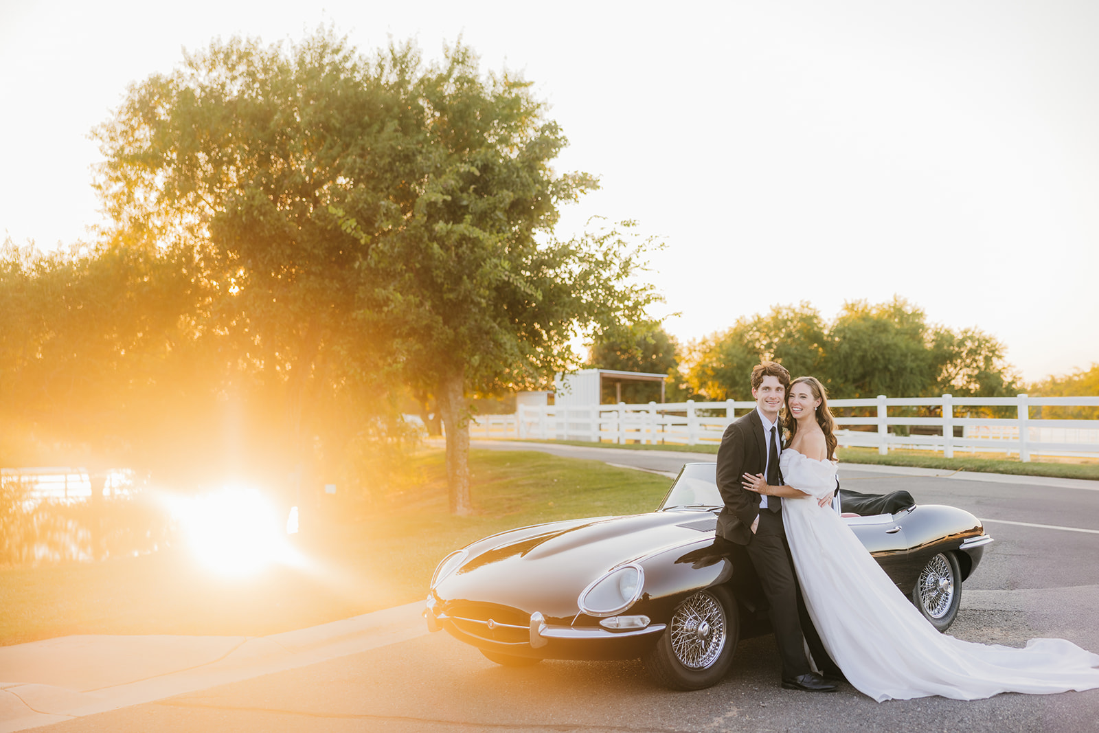 A couple in wedding attire stands by a vintage black convertible on a sunlit road, with trees and a fence in the background at aspen ranch