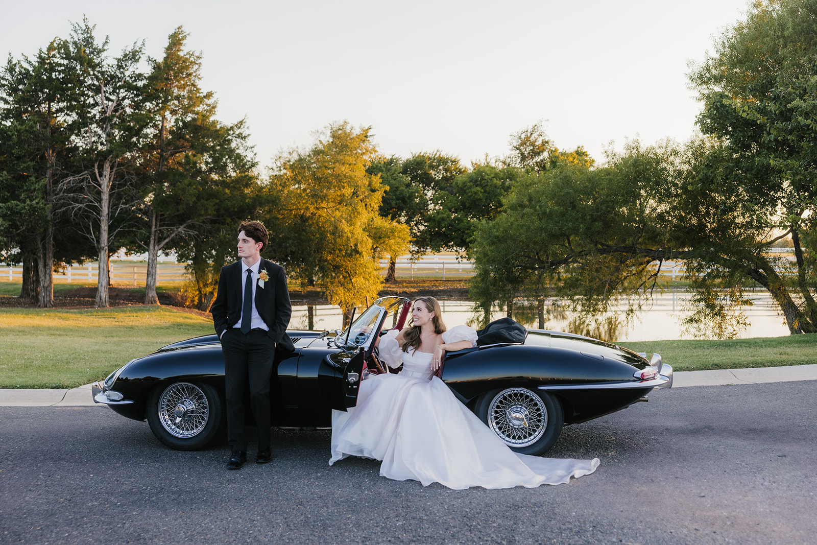 A couple in wedding attire stands by a vintage black convertible on a sunlit road, with trees and a fence in the background at aspen ranch