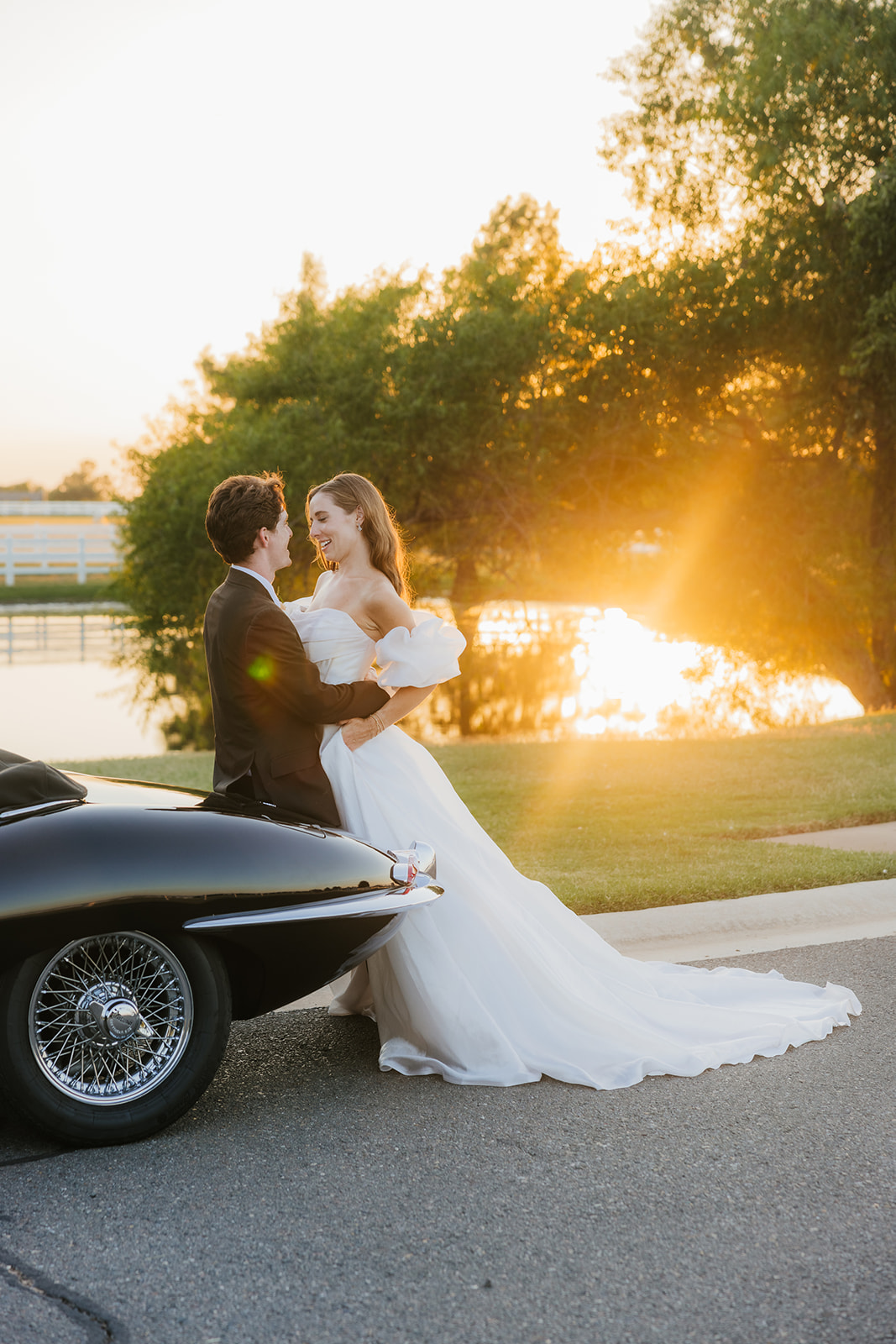 A couple in wedding attire stands by a vintage black convertible on a sunlit road, with trees and a fence in the background at aspen ranch