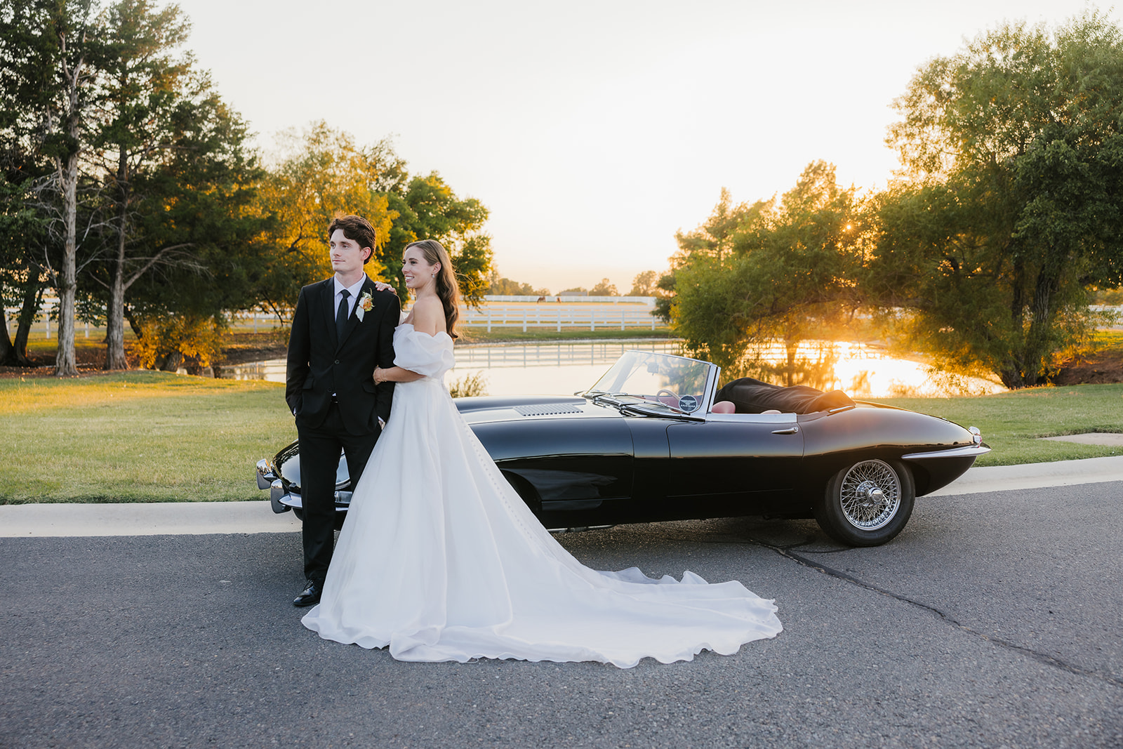 A couple in wedding attire stands by a vintage black convertible on a sunlit road, with trees and a fence in the background at aspen ranch