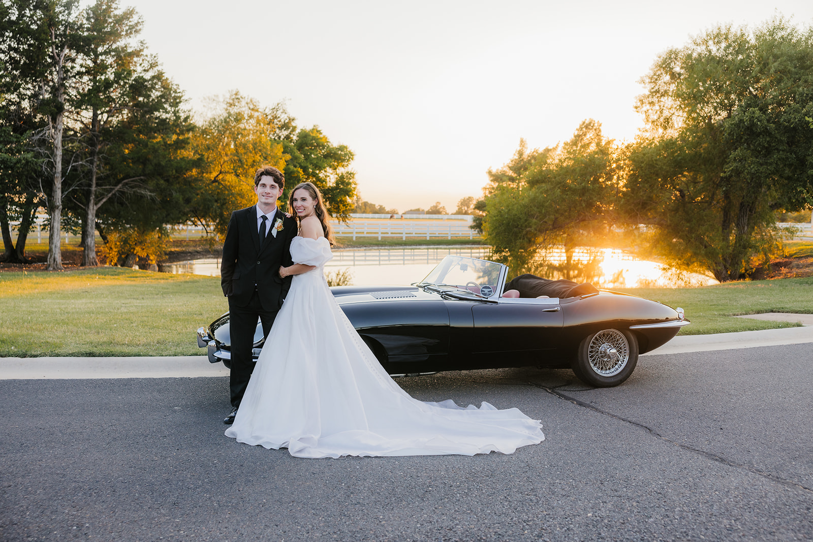 A couple in wedding attire stands by a vintage black convertible on a sunlit road, with trees and a fence in the background at aspen ranch