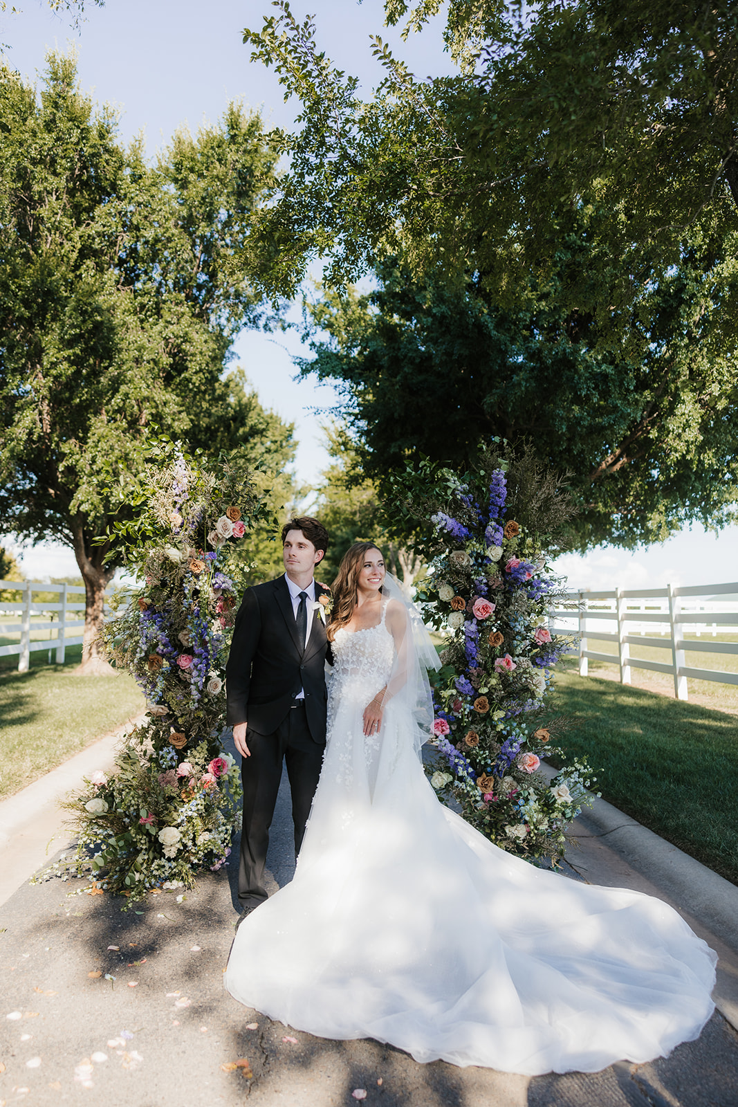 A bride and groom stand together outdoors, surrounded by two floral arrangements. The bride wears a white gown and the groom is in a dark suit. There are trees and a white fence in the background as aspen ranch