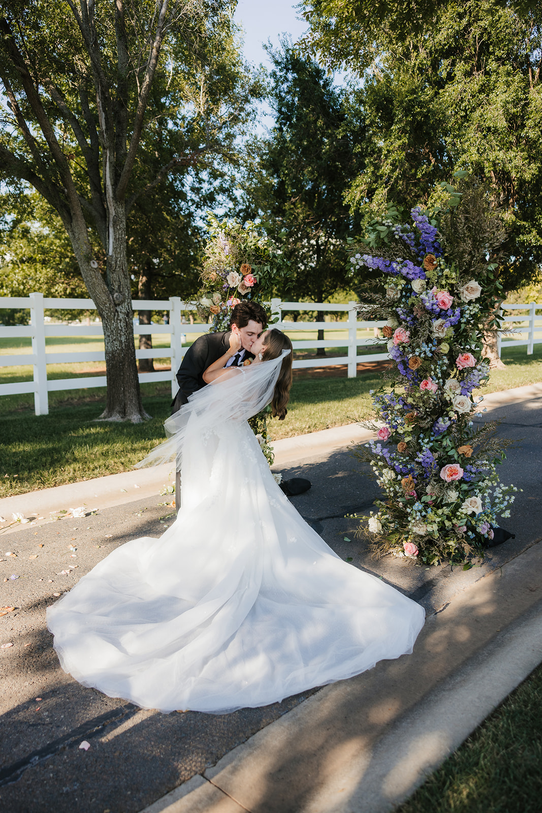 A bride and groom stand together outdoors, surrounded by two floral arrangements. The bride wears a white gown and the groom is in a dark suit. There are trees and a white fence in the background as aspen ranch