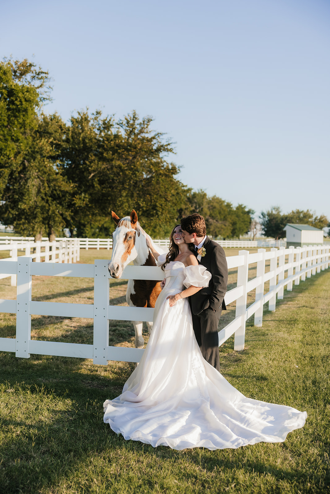 Bride and groom stand next to a white fence with a horse, in a grassy field. The bride wears a long white dress, and the groom is in a dark suit at aspen ranch