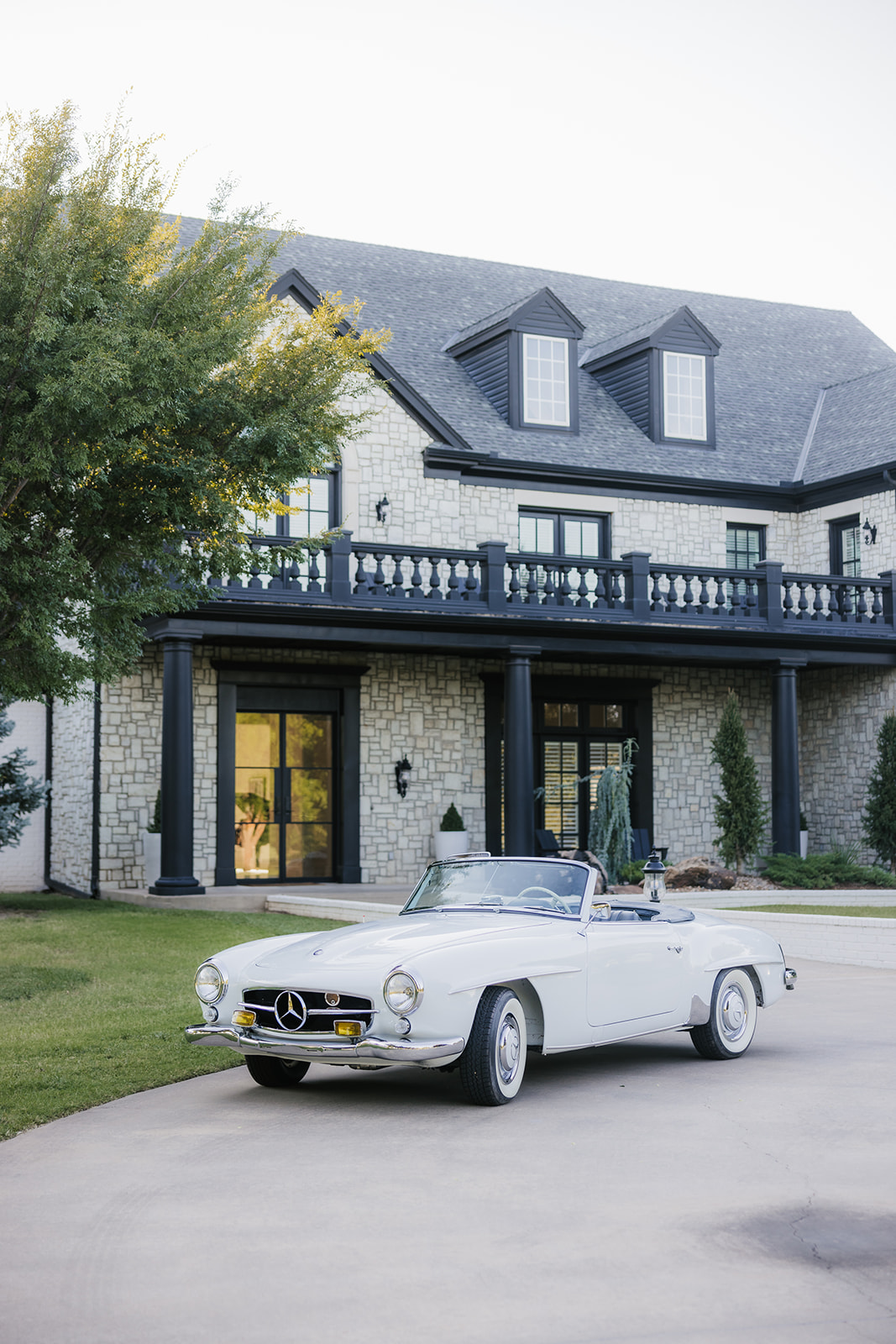 A vintage white convertible car is parked on a driveway in front of a large stone house with black trim. Trees and grass surround the area.