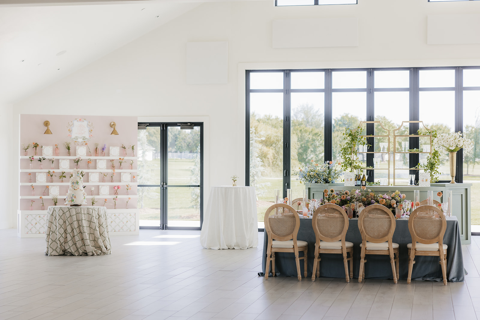 Bright reception area with a dining table set for eight, featuring elegant decorations and high-back chairs. A decorative shelf and round table are displayed against large windows in the background.