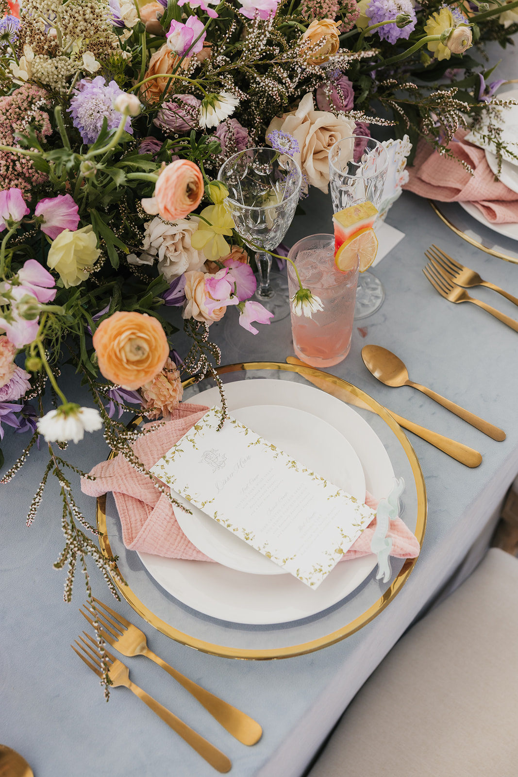 Elegant table setting with floral centerpiece, plates, gold cutlery, and a drink garnished with citrus. Napkin folded neatly beside menu card on blue tablecloth as aspen ranch