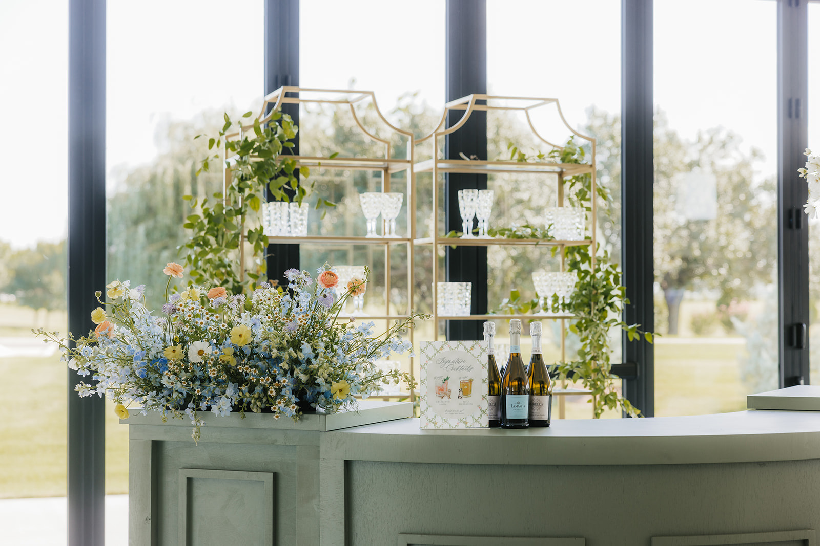 Elegant bar setup with glass shelves, floral arrangements, and bottles of drinks in front of large windows with a view of trees.