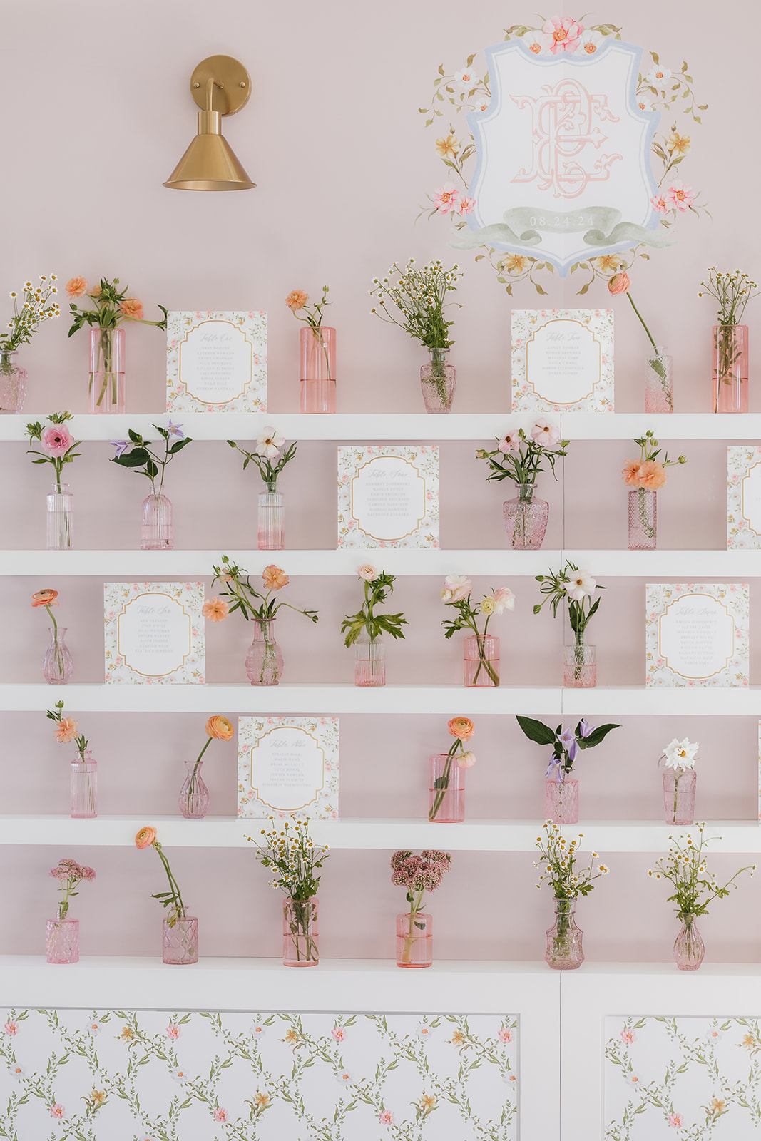 Wall-mounted shelf with small pink vases holding assorted flowers. Decorative plates are displayed alongside the vases. A light fixture is above, with floral patterns on the wall and lower panel.