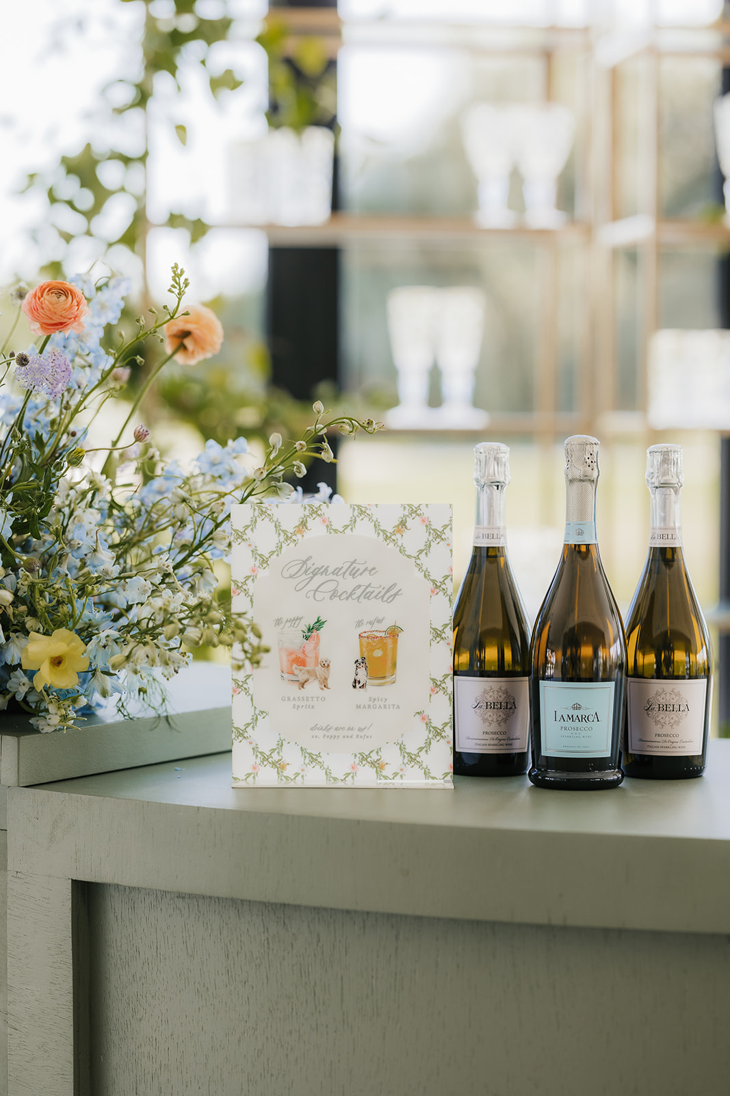 Three bottles of sparkling wine and a floral arrangement on a bar counter with a decorative sign displaying drink illustrations.