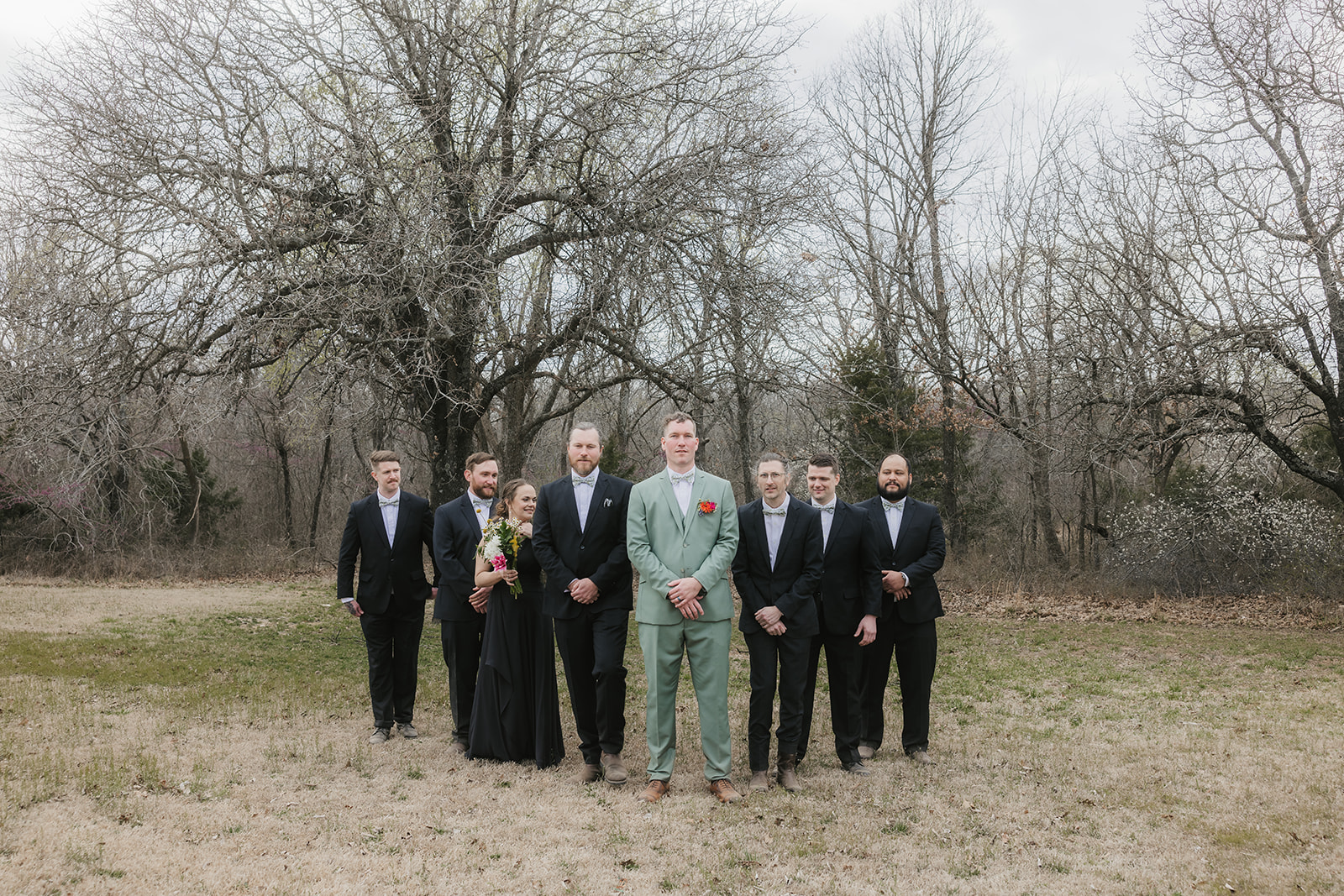 Eight people dressed formally, including one in a light green suit, stand outdoors on grass with bare trees in the background, posing for a group photo.