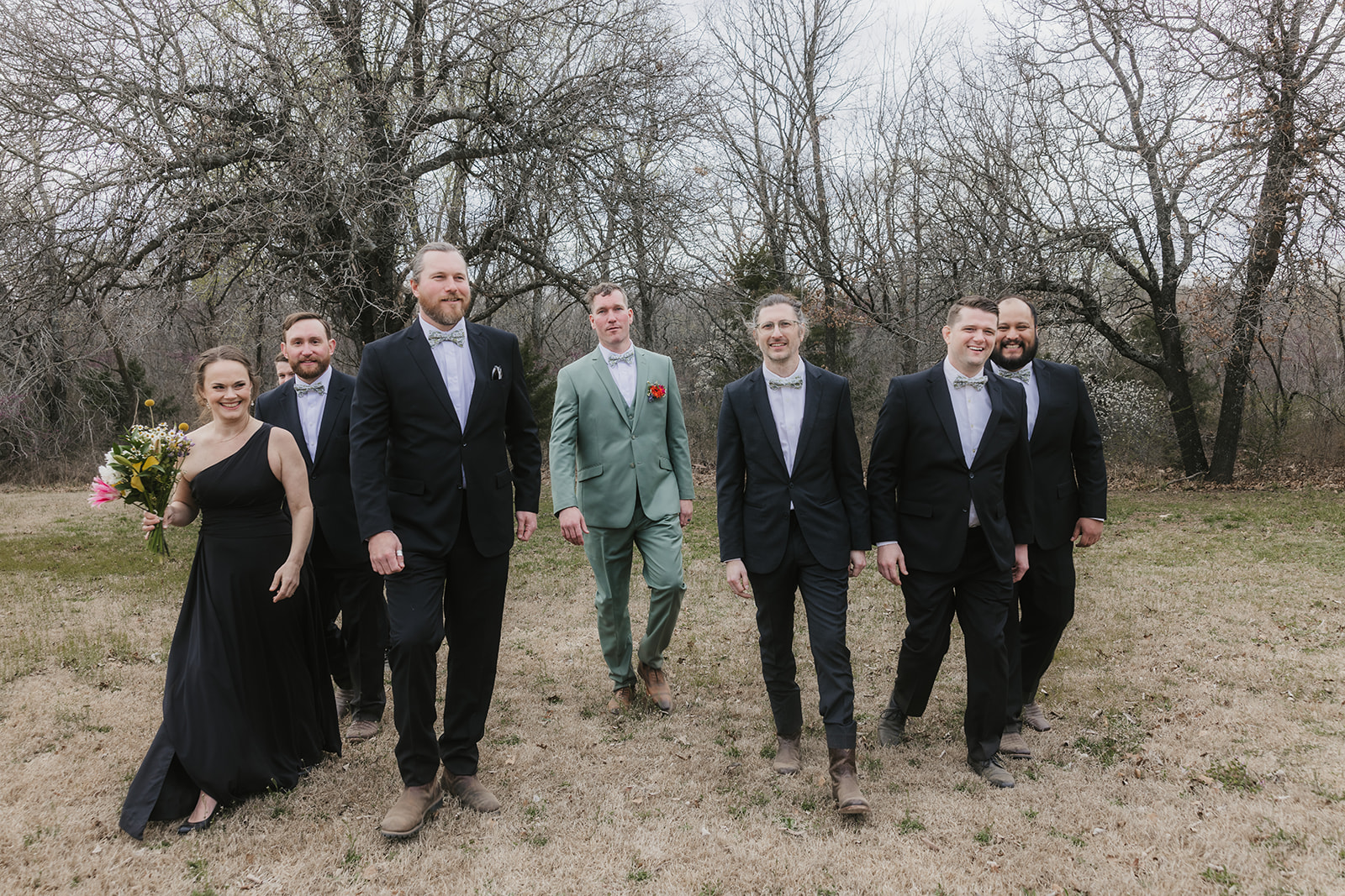 Eight people dressed formally, including one in a light green suit, stand outdoors on grass with bare trees in the background, posing for a group photo.