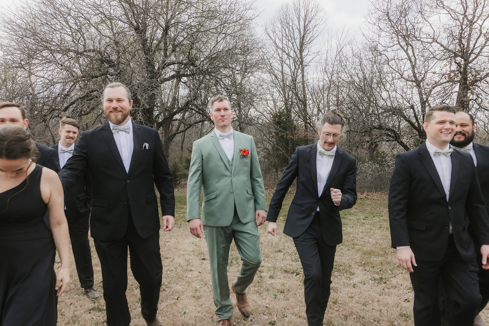 Eight people dressed formally, including one in a light green suit, stand outdoors on grass with bare trees in the background, posing for a group photo.