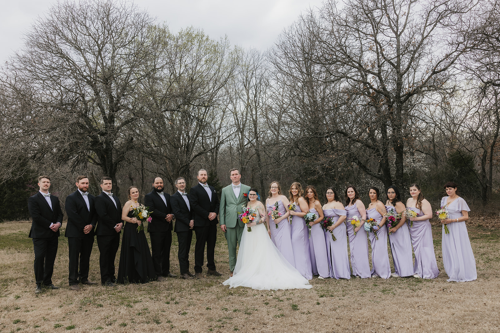 A wedding party poses outdoors in formal attire, with the bride and groom in the center, bridesmaids in light lavender dresses, and groomsmen in dark suits.