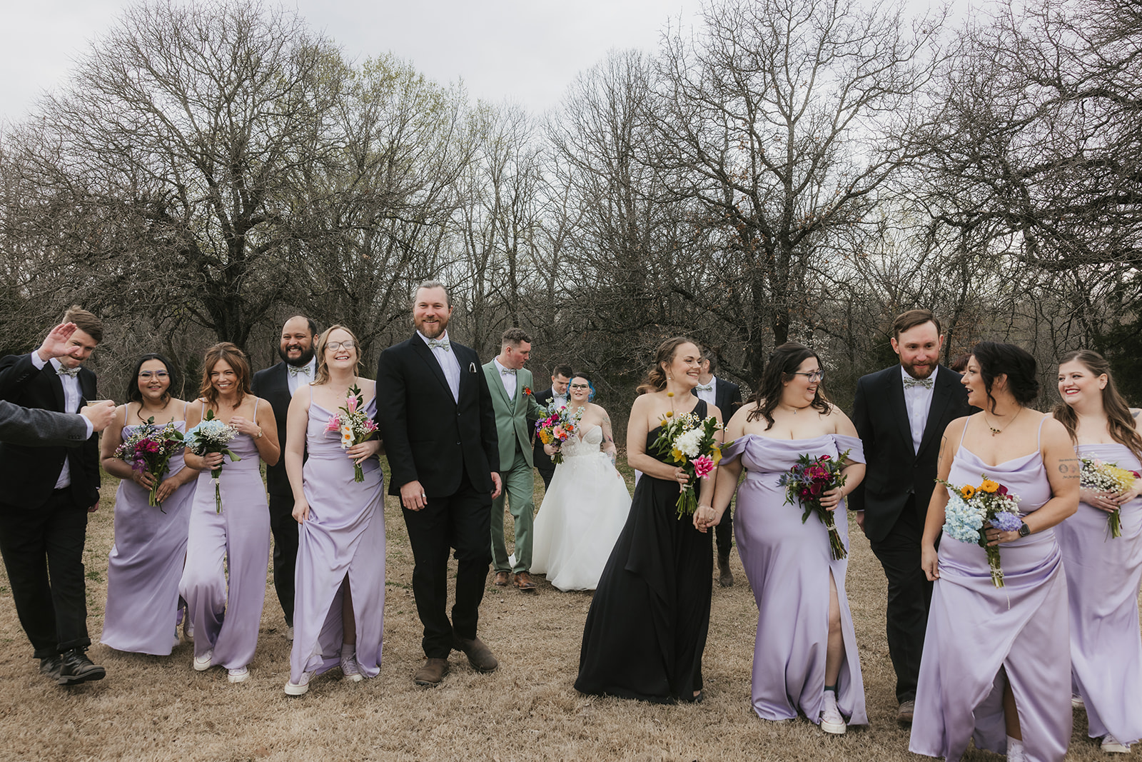 A wedding party poses outdoors in formal attire, with the bride and groom in the center, bridesmaids in light lavender dresses, and groomsmen in dark suits.