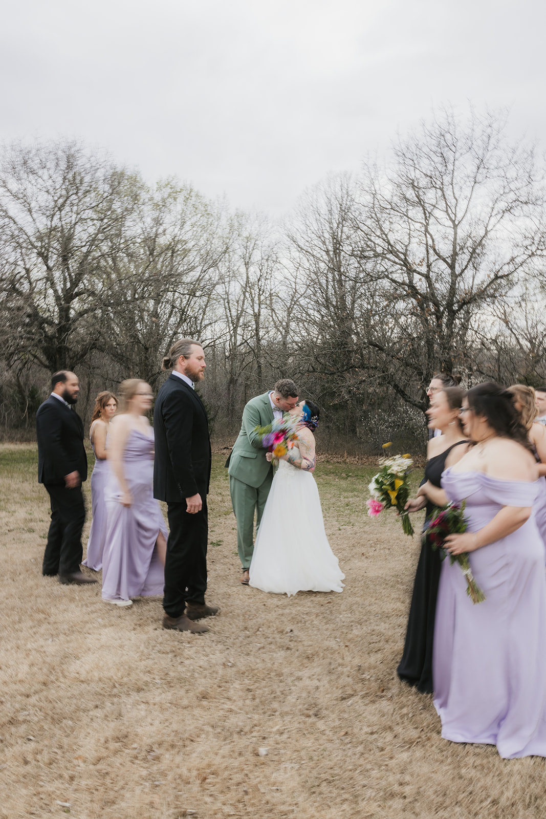 A wedding party poses outdoors in formal attire, with the bride and groom in the center, bridesmaids in light lavender dresses, and groomsmen in dark suits.