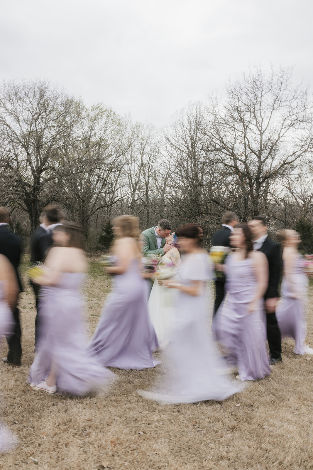 A wedding party poses outdoors in formal attire, with the bride and groom in the center, bridesmaids in light lavender dresses, and groomsmen in dark suits.