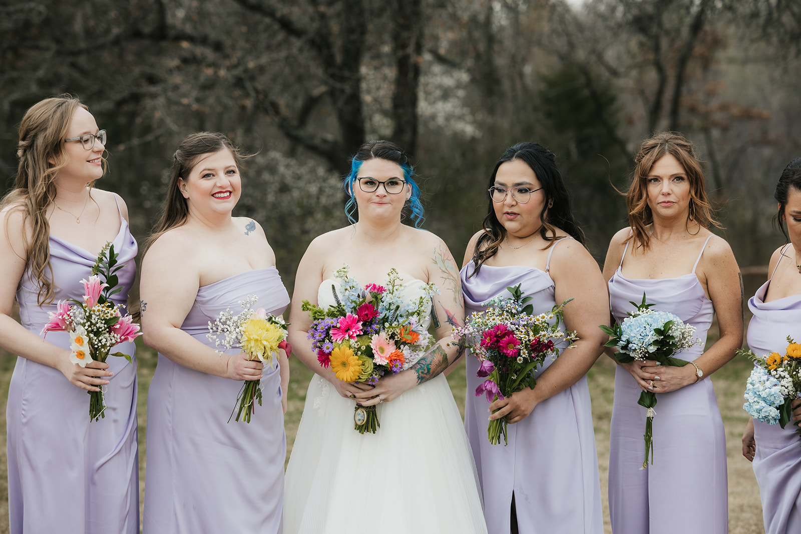 A wedding party poses outdoors in formal attire, with the bride and groom in the center, bridesmaids in light lavender dresses, and groomsmen in dark suits.
