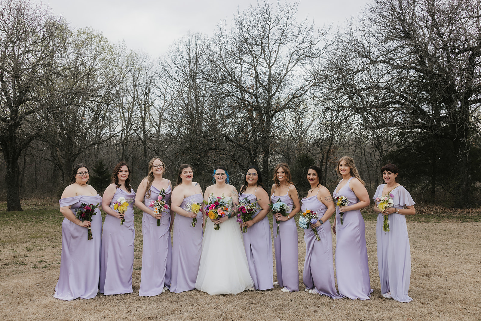 A wedding party poses outdoors in formal attire, with the bride and groom in the center, bridesmaids in light lavender dresses, and groomsmen in dark suits.