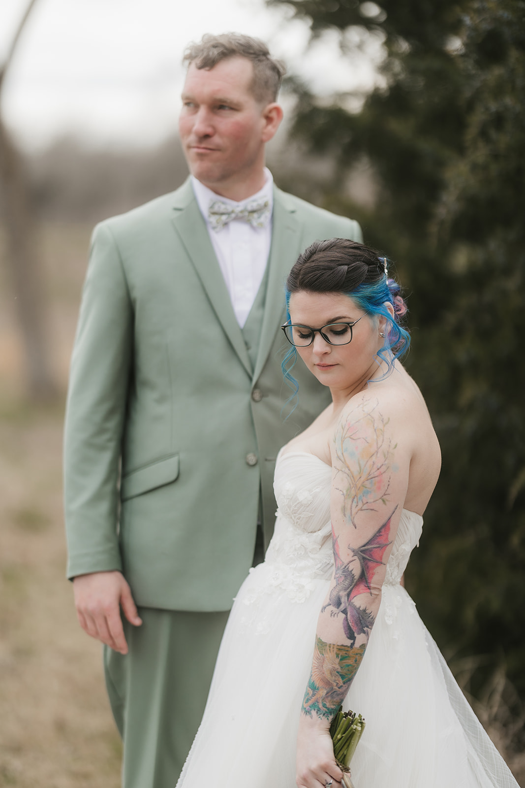 A couple in wedding attire stands outdoors; the groom leans in to kiss the bride, who holds a colorful bouquet. Leafless trees and sparse grass are in the background for a wedding at Discoveryland Ranch