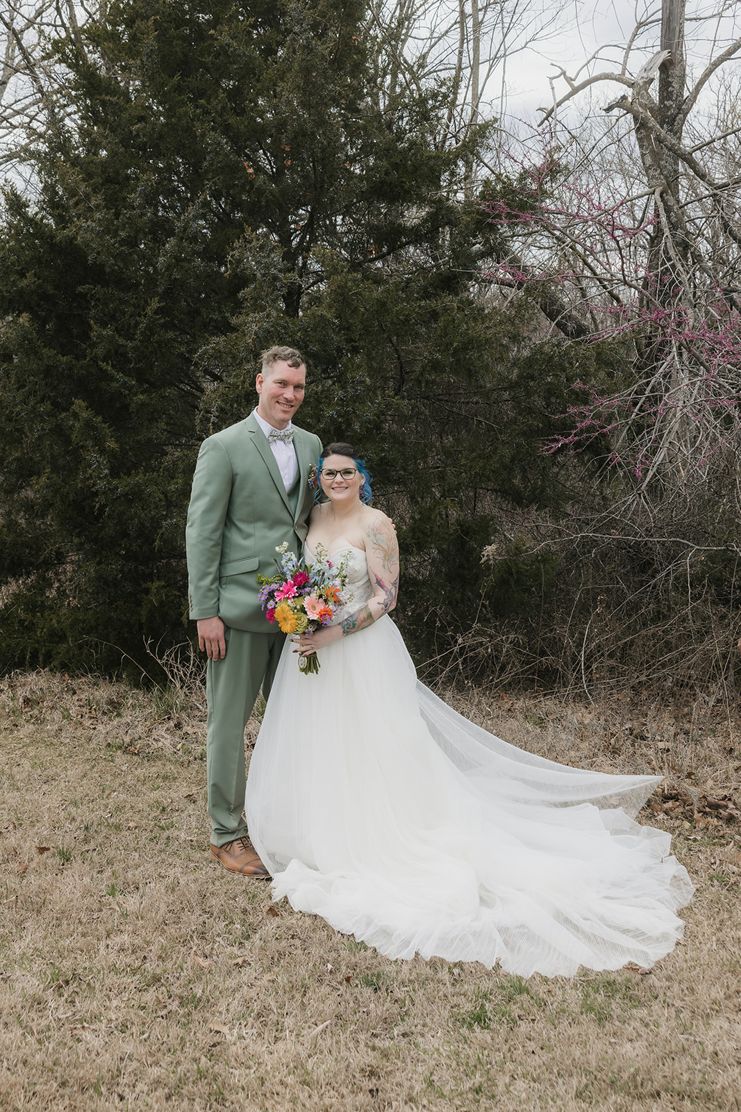 A couple in wedding attire stands outdoors; the groom leans in to kiss the bride, who holds a colorful bouquet. Leafless trees and sparse grass are in the background for a wedding at Discoveryland Ranch