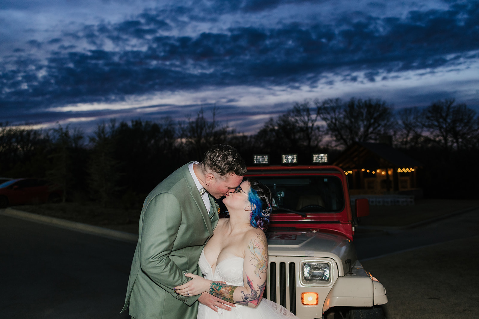 A couple dressed in wedding attire kisses in front of a red Jeep at dusk, with a dark, cloudy sky and trees in the background.