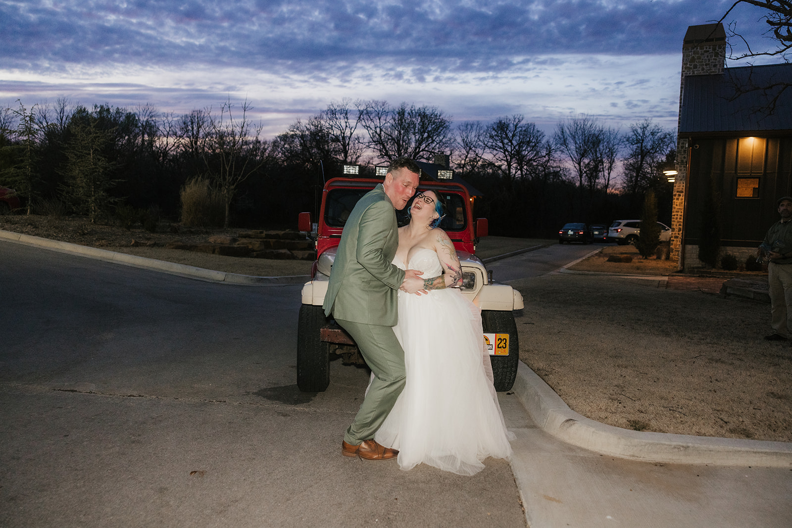 A couple dressed in wedding attire kisses in front of a red Jeep at dusk, with a dark, cloudy sky and trees in the background.