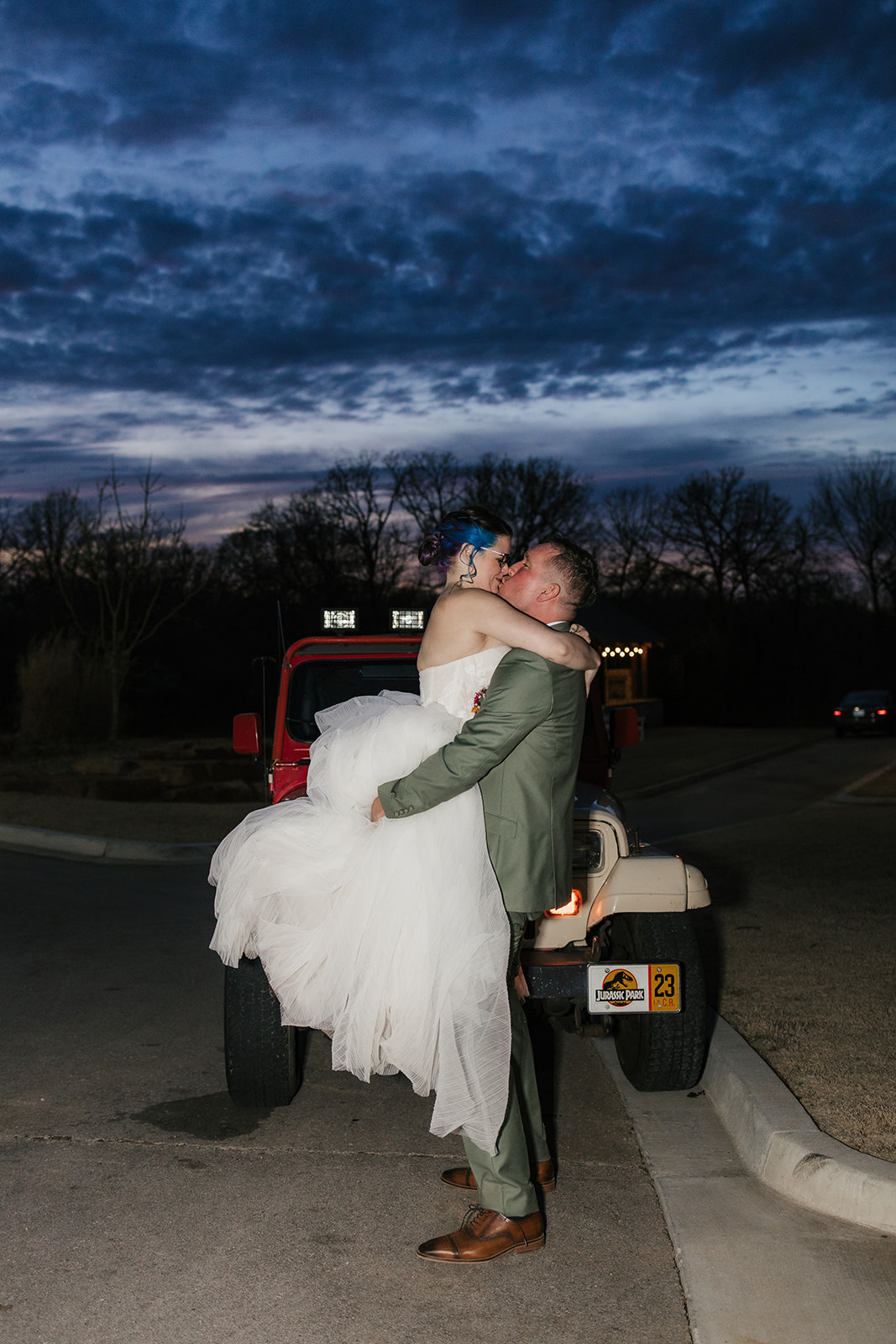 A couple dressed in wedding attire kisses in front of a red Jeep at dusk, with a dark, cloudy sky and trees in the background.