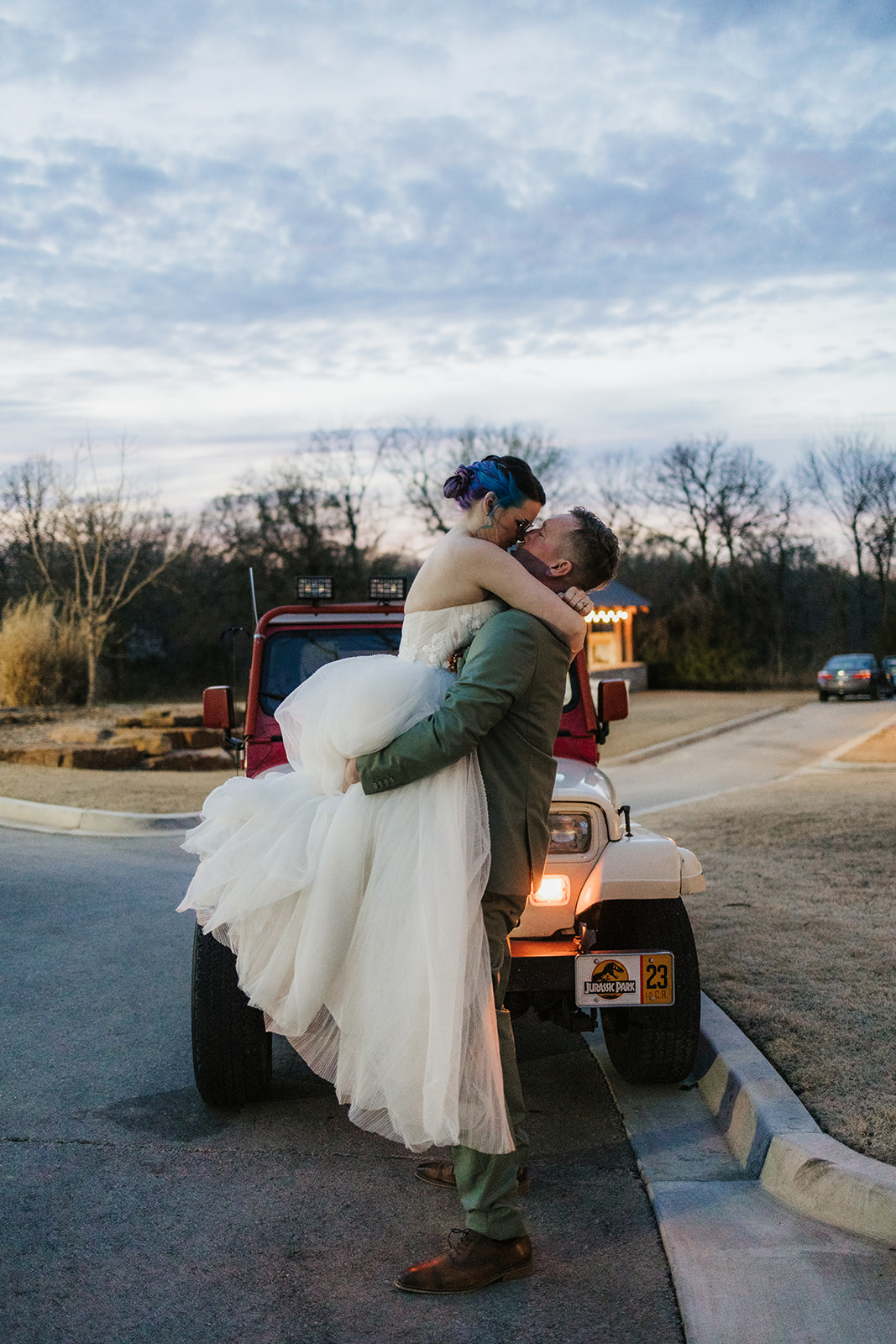 A couple dressed in wedding attire kisses in front of a red Jeep at dusk, with a dark, cloudy sky and trees in the background.