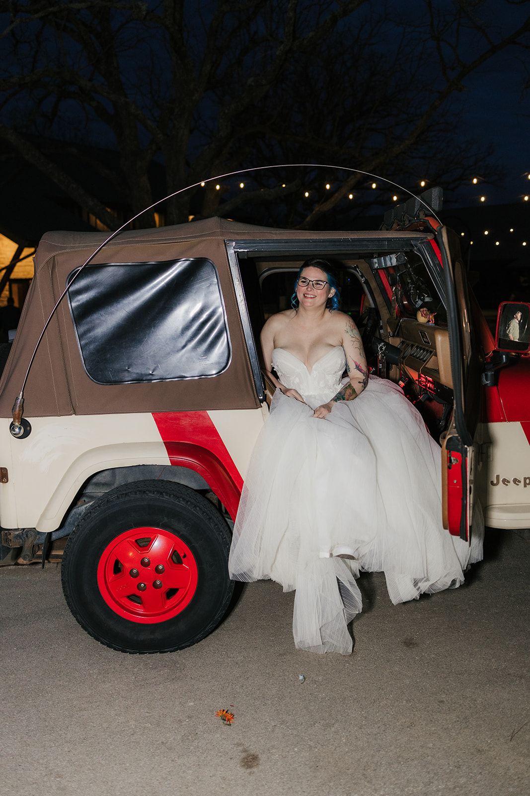 A couple dressed in wedding attire kisses in front of a red Jeep at dusk, with a dark, cloudy sky and trees in the background.