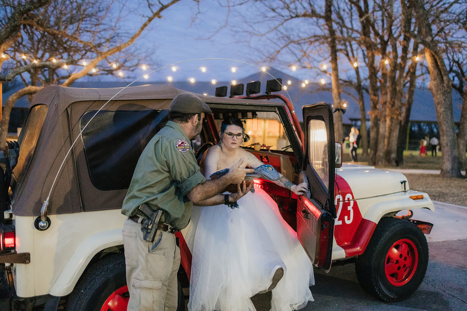 A couple dressed in wedding attire kisses in front of a red Jeep at dusk, with a dark, cloudy sky and trees in the background.