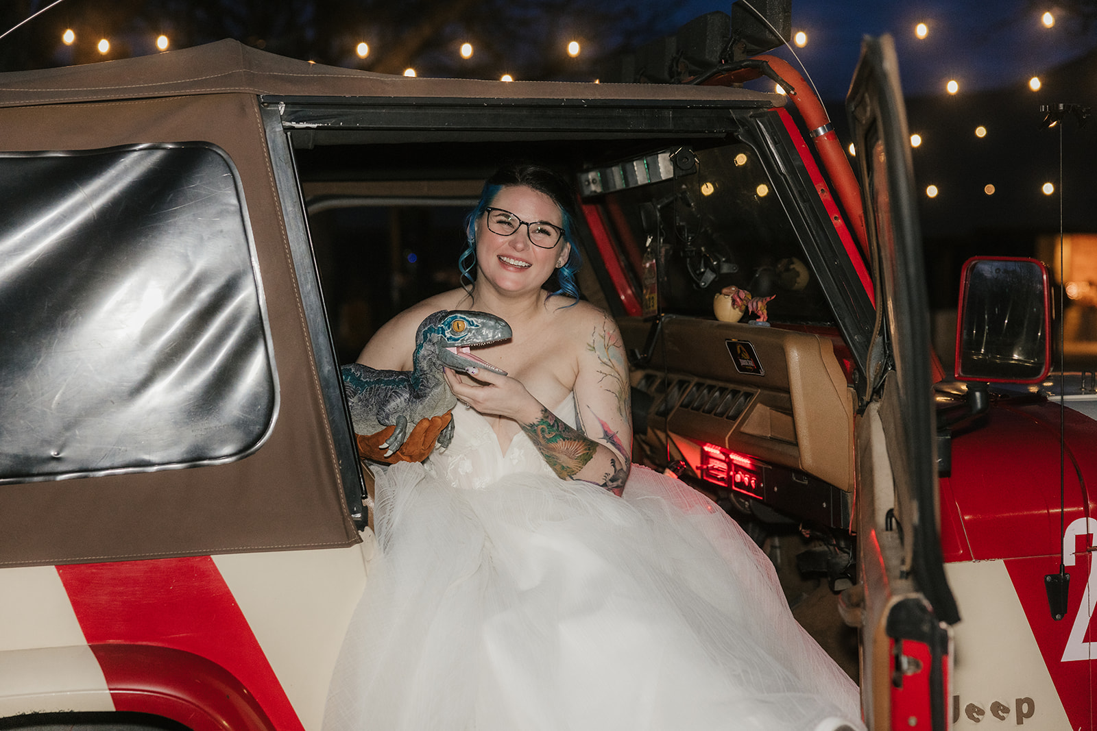 A couple dressed in wedding attire kisses in front of a red Jeep at dusk, with a dark, cloudy sky and trees in the background.