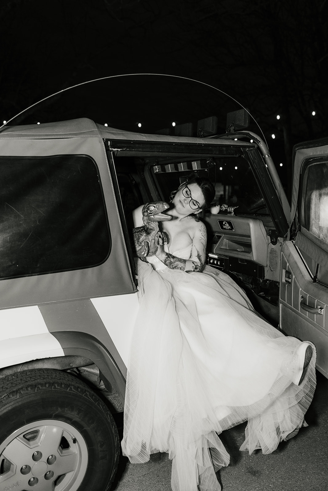 A couple dressed in wedding attire kisses in front of a red Jeep at dusk, with a dark, cloudy sky and trees in the background.