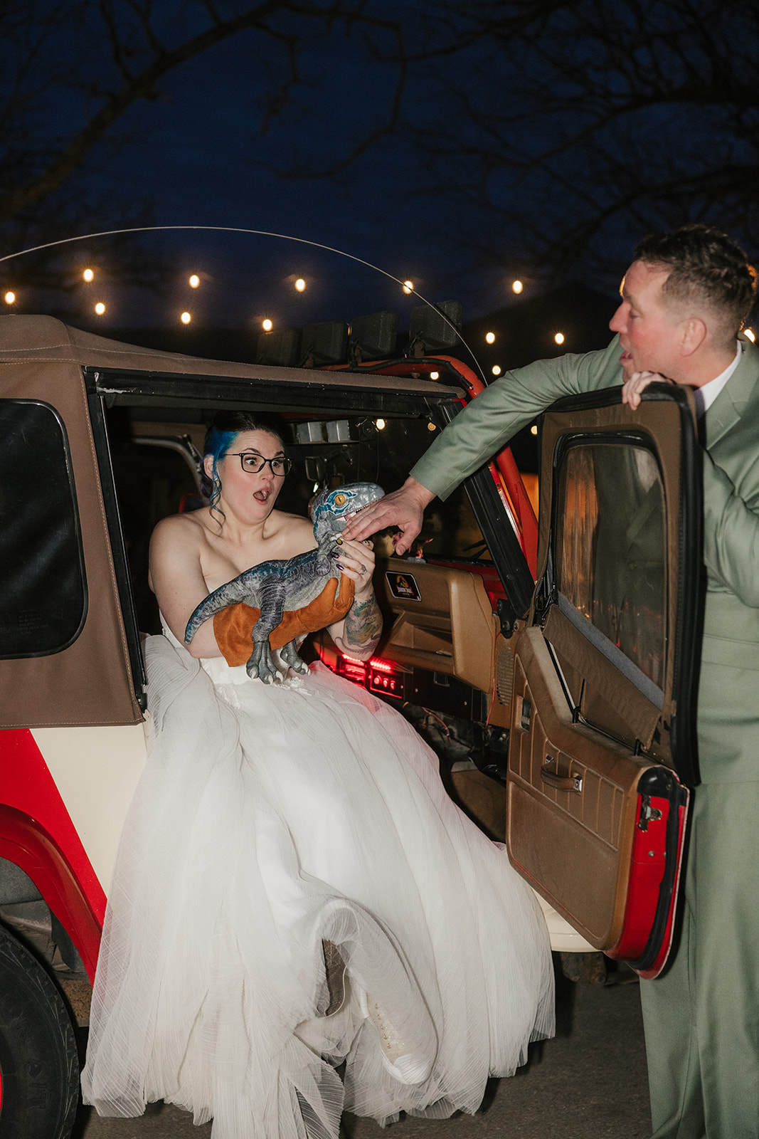 A couple dressed in wedding attire kisses in front of a red Jeep at dusk, with a dark, cloudy sky and trees in the background.