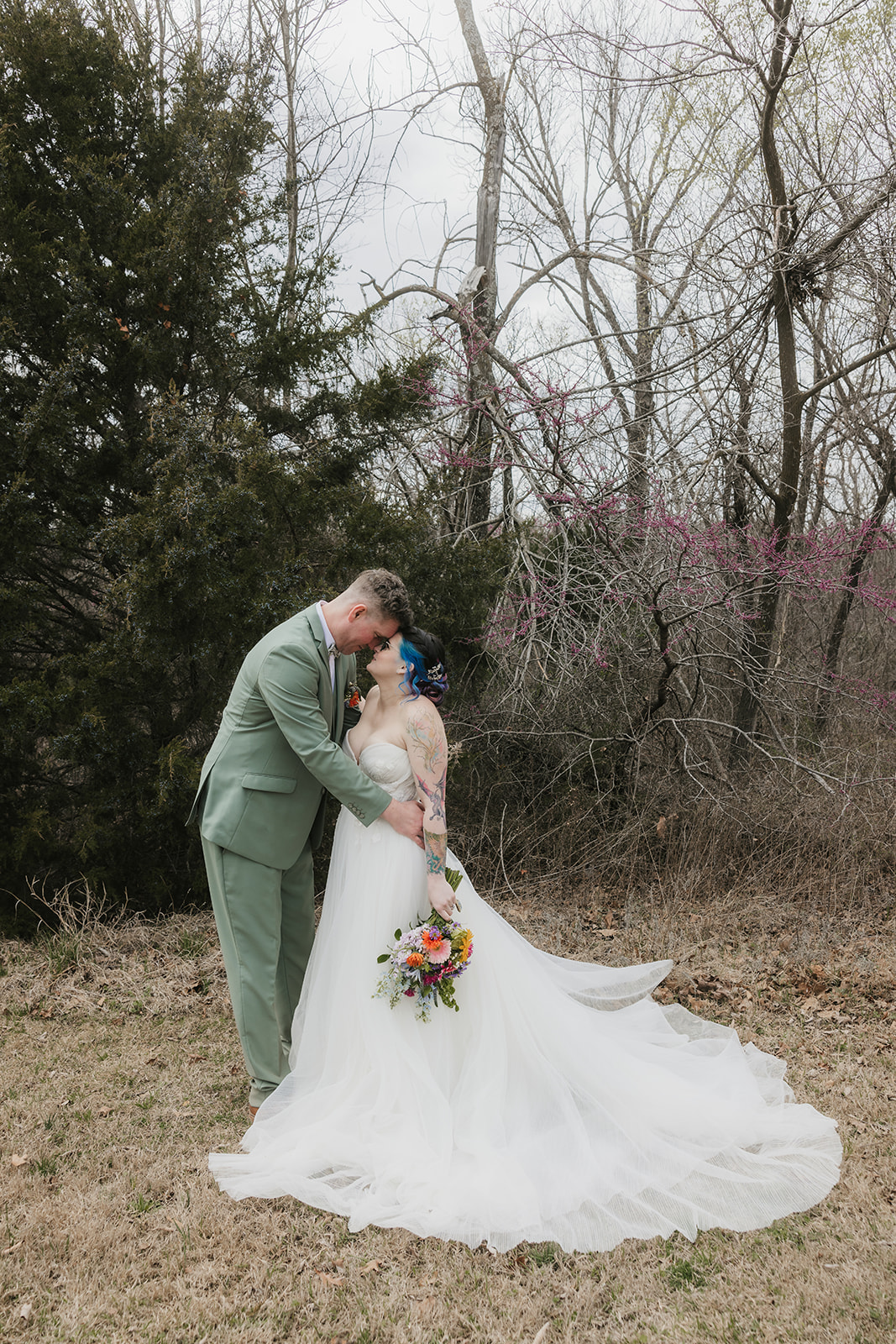 A couple in wedding attire stands outdoors; the groom leans in to kiss the bride, who holds a colorful bouquet. Leafless trees and sparse grass are in the background for a wedding at Discoveryland Ranch